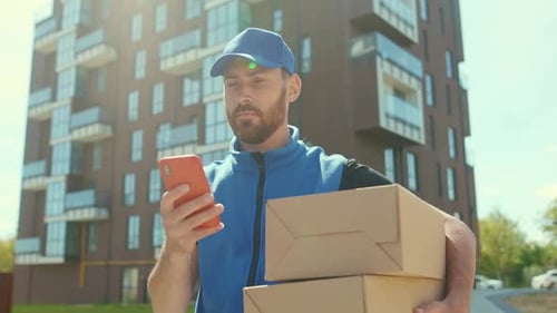 Attractive Young Man Delivery Texting Message on Smartphone Outdoor and Looking for Address Hold Box
