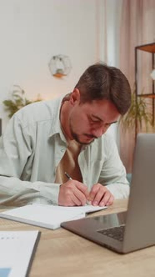 Young Man Writing Down Notes While Attending Online Office Meeting on Laptop at Home Office Desk