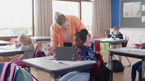 In school, female teacher helping student with tablet in classroom, other students studying