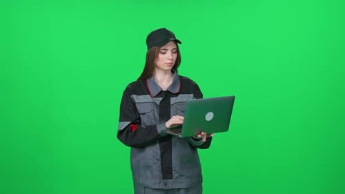 Focused Woman Worker in a Uniform Technical Engineer Stands and Uses a Laptop on a Green Background