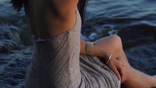 A woman in a beautiful dress sitting on a rock in the water. Female hands with bracelets dipping in