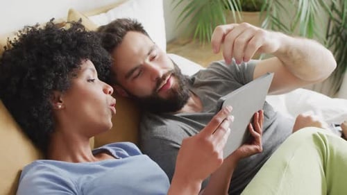 Video of happy diverse couple relaxing at home, lying on bed using tablet