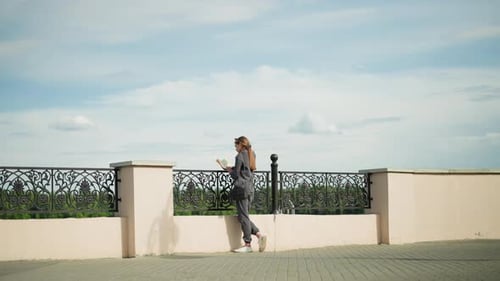 Lady in Grey Reading By Fence with Trees and Lamp Post in Background