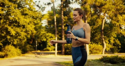 A Sports Girl in a Sports Summer Uniform is Jogging in the Morning Park and Pouring Water Over