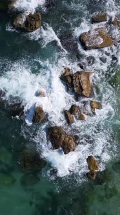 Aerial view of waves crashing over coastal rocks on a turquoise sea, with a scenic sandy beach and f