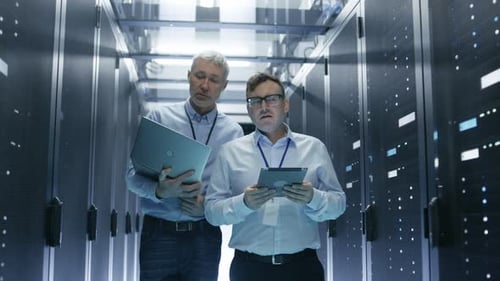 In Data Center Two IT Engineers Walking Through Rows of Server Racks. They Work on Tablet Computer