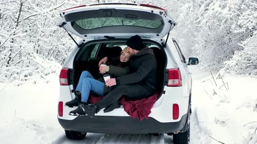 A Man and a Woman are Sitting in the Trunk of a Car in a Winter Snowy Forest Hugging
