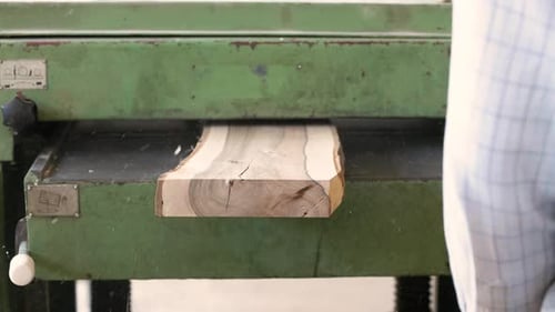 A close-up of a wooden board being processed in a green industrial planer, with sawdust flying