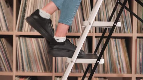 Woman Goes Up Step Ladder Near Bookcase