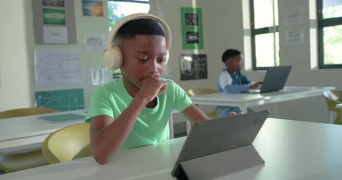 African teenage boy wearing beige headphones tapping tablet folio, following lesson at school desk