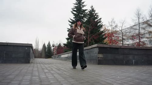 Lady Walking Slowly on Autumn Pathway Surrounded By Pine Trees and Urban Walls