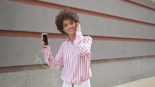 Carefree young woman dancing to music outdoors in the city with smartphone