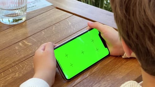 Ein Kleinkind sitzt am Holztisch und schaut mit grünem Bildschirm auf das Telefon