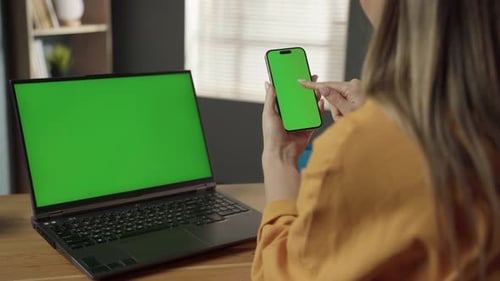 Woman Using a Smartphone with a Green Screen While a Laptop with a Green Screen Sits on a Wooden
