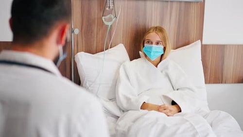 A girl in a hospital room in a medical mask holds her stomach with her hand in pain and listens