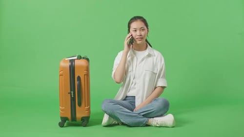 Full Body Of Female Traveler With Luggage Talking On Smartphone While Sitting In The Green Screen