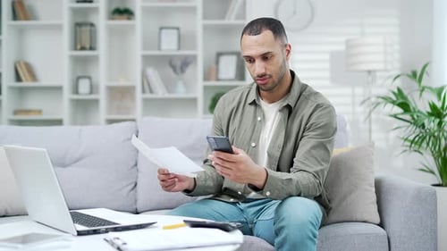 Happy man uses mobile app for spending bills using smartphone on phone while sitting at home in room