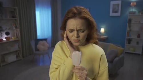 A Woman Eating Ice Cream While Standing in the Living Room The Woman Grabs Her Cheek with a