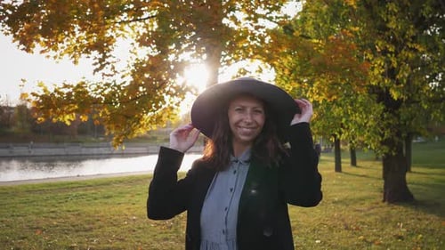 Beautiful Stylish Joyful Woman in a Black Coat and a Big Hat Walks Through the Park and Smiles