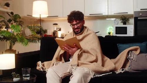 Man Reading Book on Couch Wrapped in Blanket