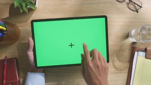 Top down view of man's hands using a green screen tablet on a wooden desk