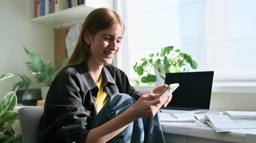 Young Cheerful Laughing Woman Reading on Smartphone Sitting at Home
