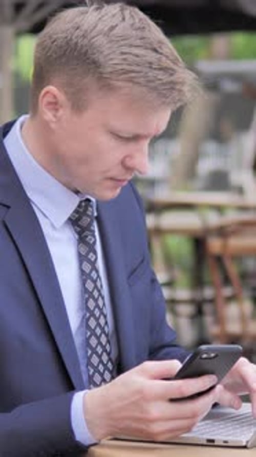 Businessman Using Smartphone and Laptop in Outdoor Cafe, Vertical Video