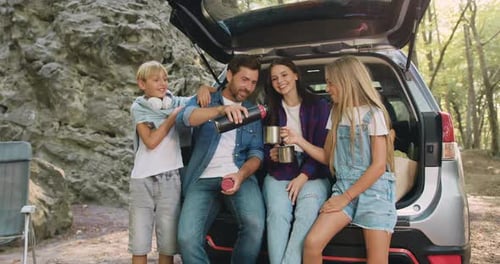 Father mother and dougher son sitting inside car trunk near rocks in forest and drinking tea