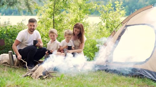 camping by the lake in the woods. happy family dad mom and little kids sitting by the fire and tent