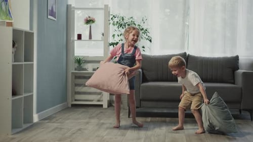 Girl Hits Younger Brother with Pillow Standing Near Sofa