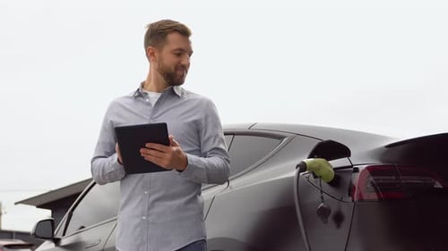 Man with Tablet Near Charging Electric Car