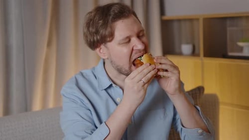 Man Sitting on Couch Eats a Burger