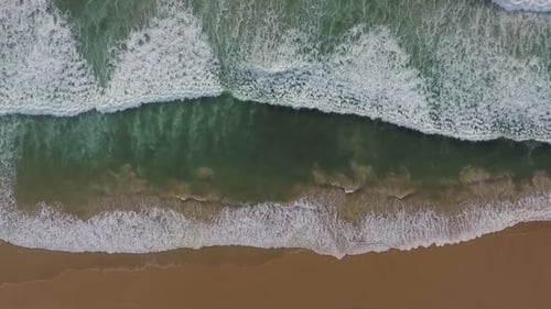 A bird's eye view of atlantic ocean waves crashing against an empty beach in slow motion