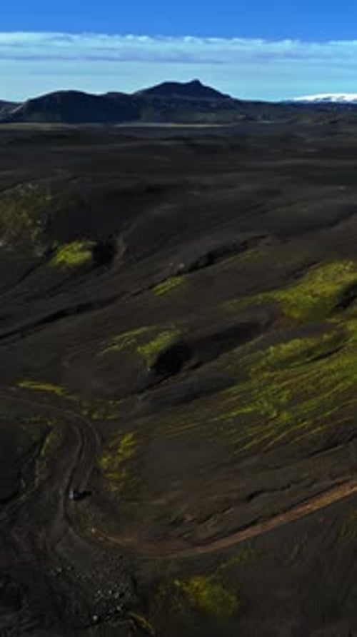Vertical drone shot of a lone SUV rounding a muddy curve through black volcanic hills streaked with