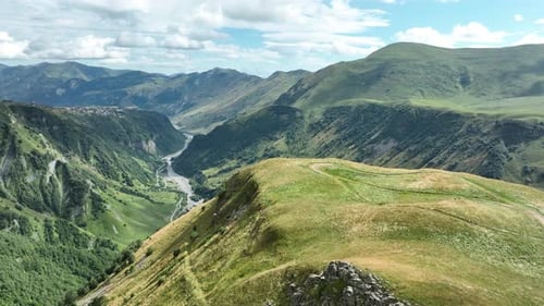 Aerial view of a deep mountain gorge with a winding riverbed, lush green slopes, and rugged peaks