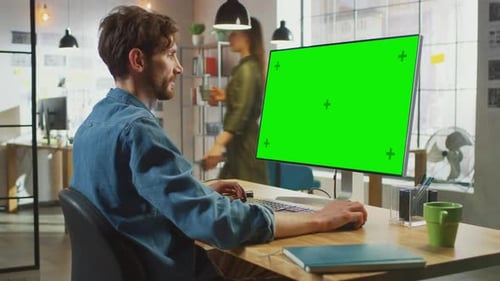Young Professional Creative Employee Works on His Personal Computer with Big Green Screen Mock Up D