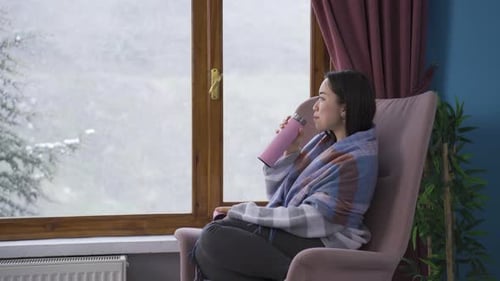 Young asian woman watching the falling snow looking out the window.