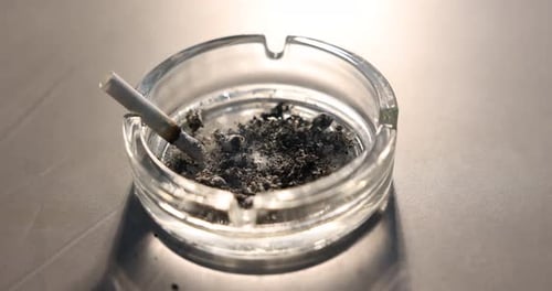 Aschenbecher mit Asche und schwelender Zigarettenkippe auf grauem Tisch, Nahaufnahme. Kamera bewegt sich vorwärts