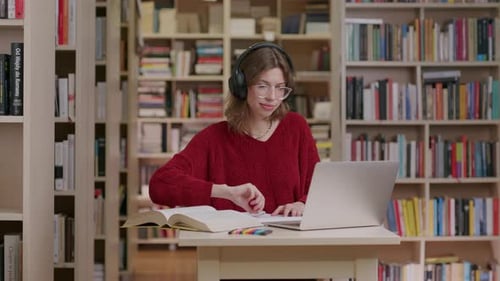 Young woman puts on headphones while studying by laptop at library