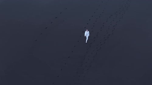 Aerial Shot of Lonely Man in White Suit Wandering on Black Sand Beach in Iceland