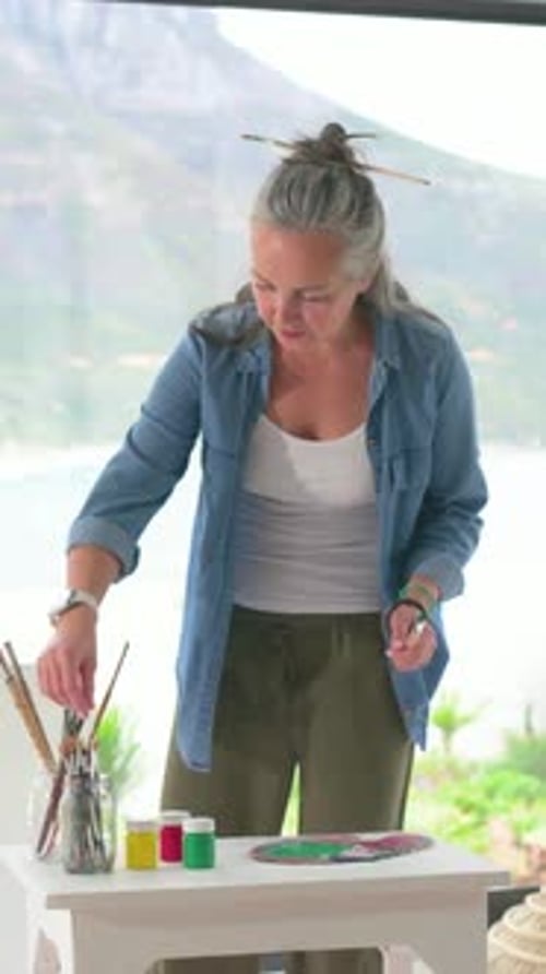 Vertical video: Starting session woman in denim mixing paint, painting by window selecting brushes
