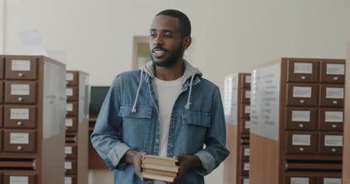 Joyful African American Man with Books in Hands Walking in Library Hall Smiling
