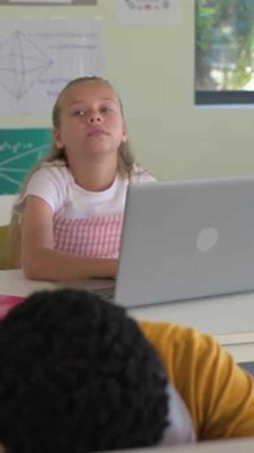 Vertical video: Camera shifting, girl in pink dress leaning in class with laptop, boy lowering head