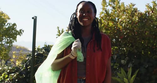 Mujer africana que lleva una bolsa verde con guantes con plástico atado durante la limpieza del césped