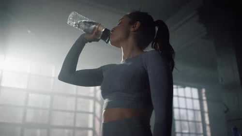 Tired Woman Drinking Water in Dark Gym. Thirsty Girl Taking Rest At