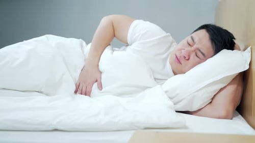 young sick asian man with severe abdominal pain, stomach ache poisoning. lying in bed in the morning