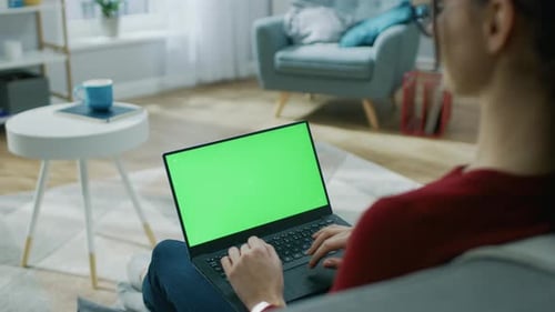 Young Woman at Home Works on a Laptop Computer with Green Mock-up Screen. He's Sitting On a Couch i