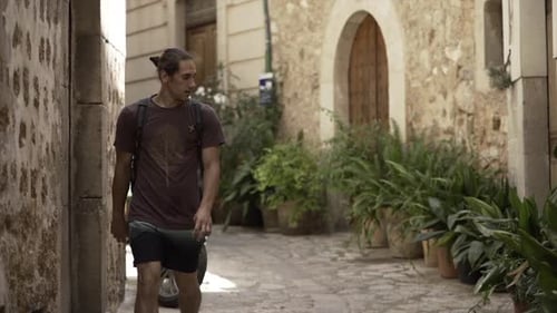 Young Athletic Boy with Long Hair Walks Down a Town Rustic Street in Summer