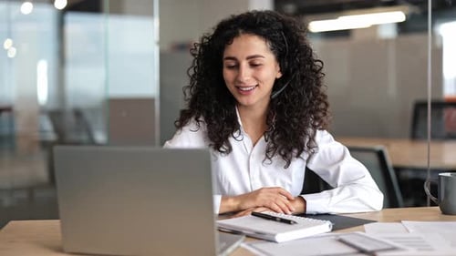 Businesswoman in Headset Smiles During Video Conference on Laptop in Office