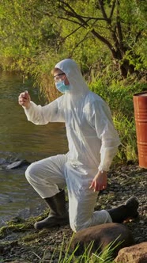 Environmental inspector in full hazmat suit collecting water samples from a sunlit river.